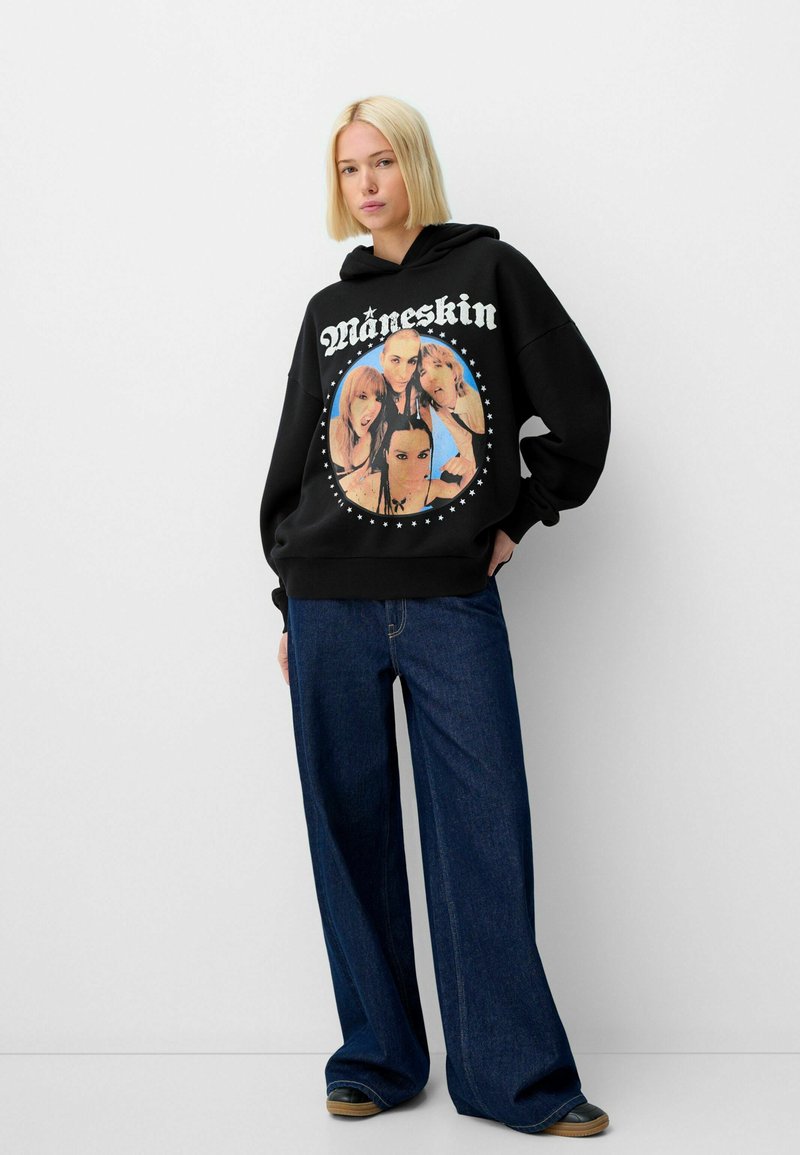 Felpa nera con cappuccio caratterizzata da una grafica circolare che presenta quattro individui circondati da stelle bianche, abbinata a jeans a gamba larga in denim scuro.