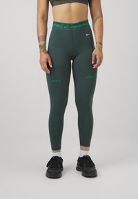 Nike Pro legíny ve tmavě zelené barvě, úzký střih, vysoký pas s zeleným logem, délka do poloviny lýtek, měkký materiál, v kombinaci s černými teniskami.