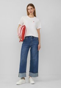 T-shirt en coton blanc avec logo en forme de cœur rouge, jeans bleu à jambes larges avec ourlet retroussé, et baskets blanches. Le mannequin tient une couverture en tricot rouge et blanche à rayures.