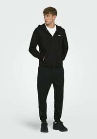 Sweat à zip noir avec cordons blancs, associé à un pantalon de jogging noir, tous deux en tissu doux ; le modèle porte également des baskets foncées.