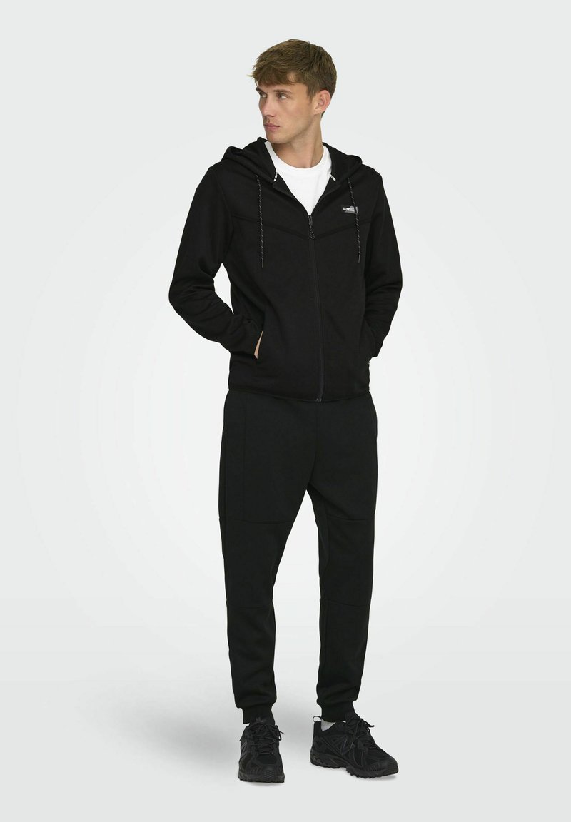 Zwarte zip-up hoodie met witte koordjes, gecombineerd met zwarte joggers, beide van zachte stof; het model draagt ook donkere sneakers.
