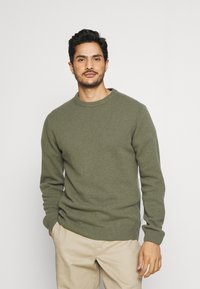 Pulover tricotat de culoare verde măsliniu, cu guler rotund, manșete și tiv cu nervuri. Confecționat dintr-un material moale, texturat. Croi casual, asortat cu pantaloni bej.