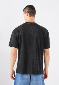 Tricou din bumbac moale, negru, cu o croială lejera, mâneci scurte, textura uzată și un decolteu rotund. Spate simplu, fără grafică vizibilă.