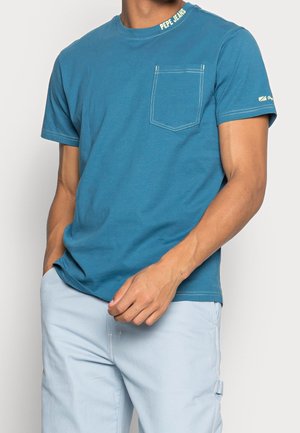 Mand iført en blå kortærmet Pepe Jeans t-shirt med brystlomme og lyseblå bukser, hånd hvilende nær linningen.