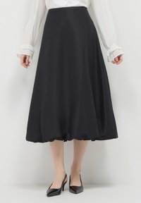 Frau trägt einen schwarzen Midirock mit einem Bubbel-Saum, eine weiße Bluse mit Rüschensäumen und schwarze Lackleder-Slingback-Pumps.