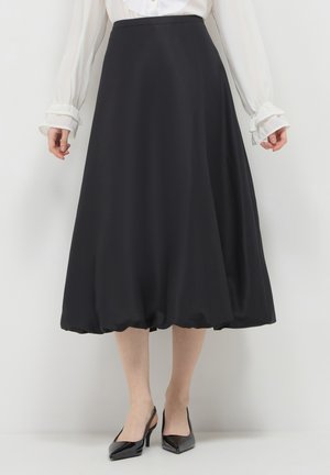 Frau trägt einen schwarzen Midirock mit einem Bubbel-Saum, eine weiße Bluse mit Rüschensäumen und schwarze Lackleder-Slingback-Pumps.