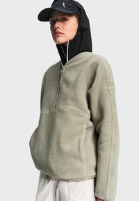 Woman wearing light green fleece pullover with half-zip and black hooded cap, standing against plain light background.