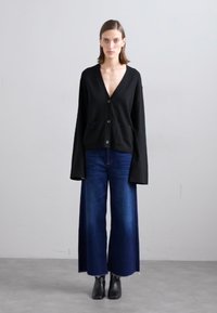 Cardigan preto de malha com decote em V, quatro botões e mangas compridas, combinado com jeans de perna larga azul escuro e botins pretos.