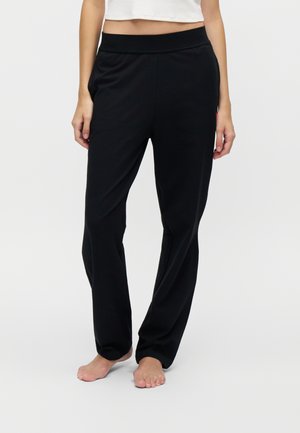 Vrouw draagt zwarte losse broek en een wit cropped topje, staat op blote voeten tegen een effen lichte achtergrond.