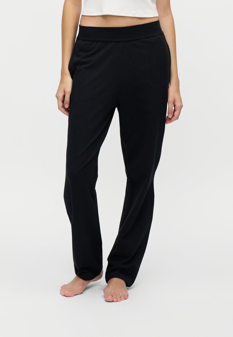 Γυναίκα με μαύρο φαρδύ παντελόνι και λευκό cropped top, ξυπόλητη, σε απλό φωτεινό φόντο.