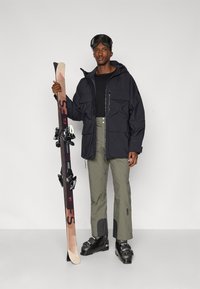Black ski jacket with large pockets, gray ski pants, and black ski boots. Holding black and beige skis with bindings. Ski goggles on head.