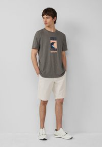 Graues Baumwoll-T-Shirt mit Rundhalsausschnitt und einem grafischen Druck, der eine Welle zeigt. Kombiniert mit cremefarbenen Shorts und weißen Sneakers.