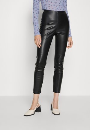 Kobieta ubrana w czarne, wysokie spodnie z ekologicznej skóry, jasnoniebieską, kwiatową bluzkę z długim rękawem oraz beżowe sandały na szerokim obcasie z paskiem z tyłu.