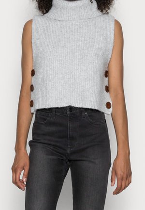 Pull sans manches en tricot gris à col roulé avec trois boutons marron de chaque côté, porté avec un jean noir taille haute.