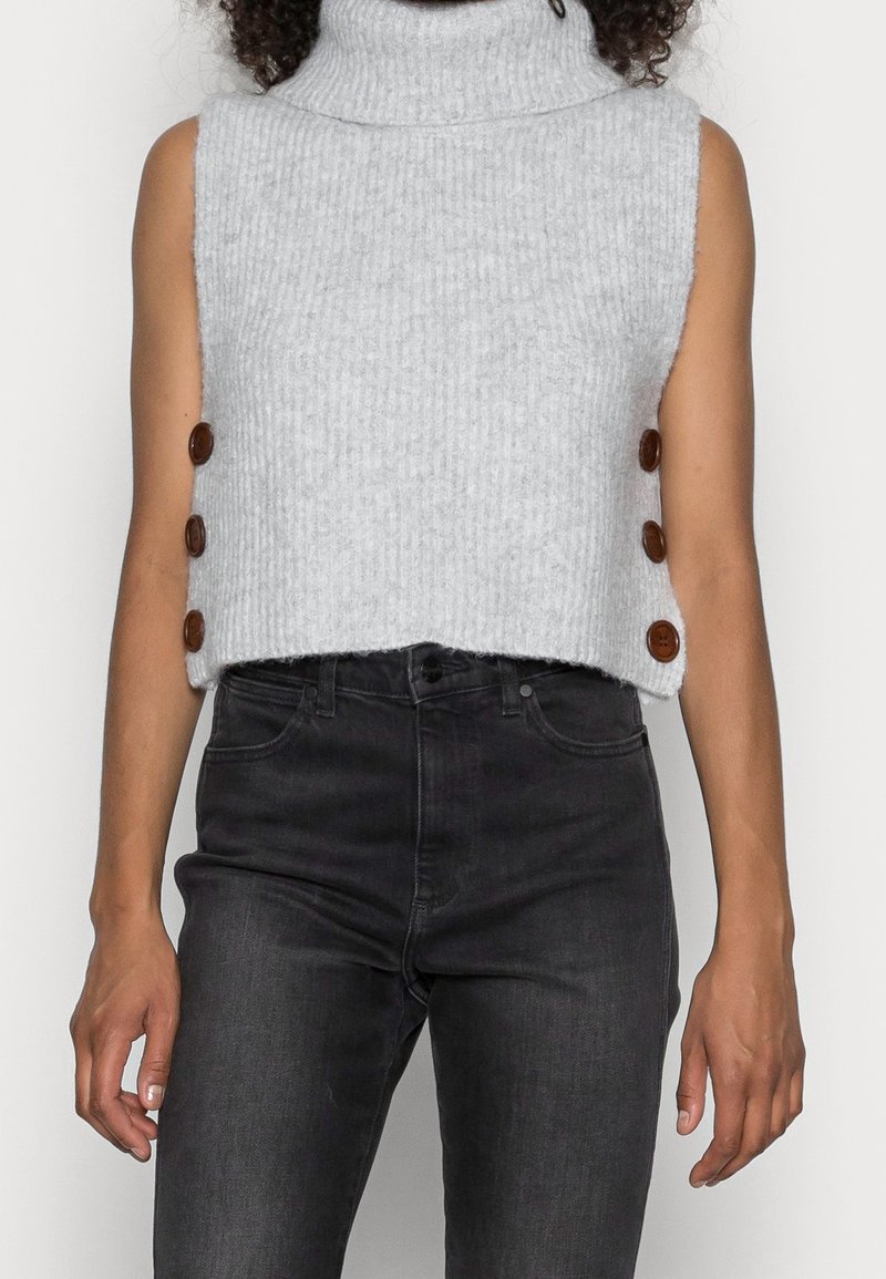 Pull sans manches en tricot gris à col roulé avec trois boutons marron de chaque côté, porté avec un jean noir taille haute.