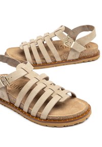 Beige Wildledersandalen mit mehreren überkreuzten Riemen, verstellbarem Knöchelverschluss, gepolstertem Fußbett und strukturiertem Gummisohle.
