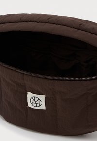 Σκούρο καφέ ύφασμα τσάντα με φερμουάρ ελαφρώς ανοιχτό, αποκαλύπτοντας ένα ευρύχωρο εσωτερικό και μια λευκή παστέλ ραμμένη στο μπροστινό μέρος.