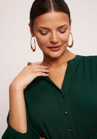 Zielona bluzka na guziki o gładkiej fakturze, z krótkimi rękawami i dekoltem w kształcie litery V. Złote kolczyki w kształcie kół i pierścionek podkreślają stylizację.