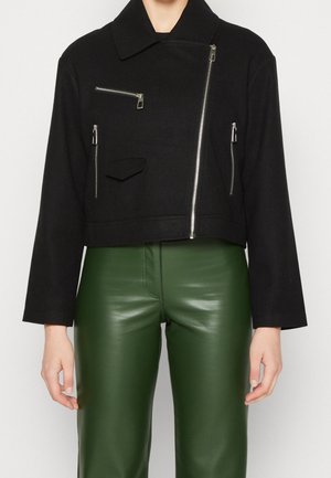 Veste courte noire avec des fermetures éclair asymétriques argentées et trois poches, portée avec un pantalon en cuir vert taille haute.