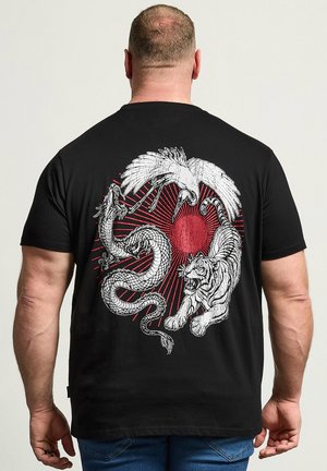 Mand iført sort skjorte med hvid drage, trane og tiger, der cirkler omkring en rød sol på ryggen, står mod en ensfarvet baggrund.