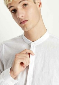 Camicia bianca in lino con colletto rigido, caratterizzata da un dettaglio con bottone alla scollatura e un logo ricamato discreto sul tessuto.