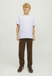 Blondais zēns stāv, valkājot parastu baltu T-kreklu, brūnas bikses un brūnas sporta kurpes, skatās nedaudz augšup uz savu kreiso pusi.