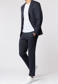 Tmavě námořnický oblek s přizpůsobeným sakem a slim-fit kalhotami. V kombinaci s bílým tričkem a bílými teniskami. Diskrétní červený akcent na saku.