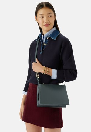 Femme aux longs cheveux noirs portant un pull bleu marine avec un col en denim, une jupe bordeaux, des bracelets dorés, et portant un sac à bandoulière en cuir vert foncé.