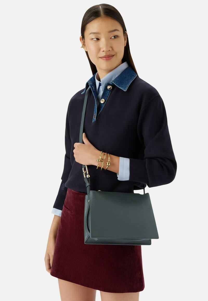 Donna con lunghi capelli scuri che indossa un maglione blu navy con colletto in denim, una gonna bordeaux e braccialetti dorati, mentre porta una borsa a spalla in pelle verde scuro.