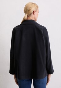 Blondide juuksed madalasse hobusesaba seotud naine, kes kannab musta jakk ja siniseid teksaseid, nähtud seljaga lihtsa valge seina taustal.