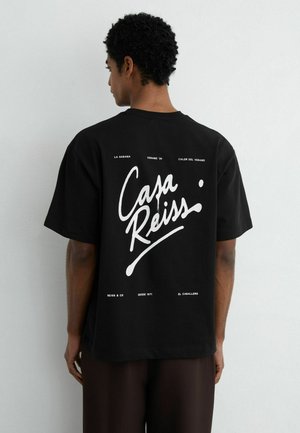 Homme aux cheveux courts et bouclés portant un T-shirt noir oversized avec un grand texte blanc "Casa Reiss" dans le dos.
