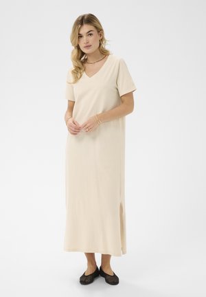 Ženska z blond lasmi, oblečena v bež midi obleko z V-izrezom, kratkimi rokavi in stranskim razporkom, ter črnimi tkanimi balerinkami, stoji z rokami sklenjenimi skupaj.