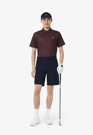Polo a maniche corte color bordeaux con un motivo a pois, abbinato a pantaloni corti blu navy. Scarpe bianche e un visiera blu; tiene in mano un bastone da golf.