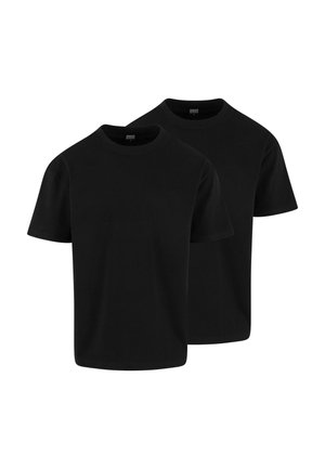 Jeune homme aux cheveux courts, debout, portant un t-shirt noir uni et un short gris clair sur un fond neutre.
