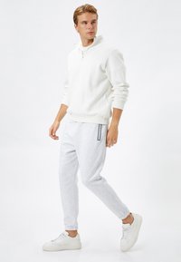 Biała bluza z suwakiem i wysokim kołnierzem, w połączeniu z jasnoszarymi spodniami dresowymi z bocznymi akcentami. Model ma na sobie białe trampki.