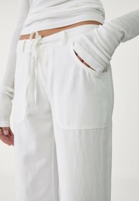 Białe, luźne spodnie wykonane z miękkiego materiału, z dużymi przednimi kieszeniami i wiązaniem w talii. Teksturowane długie rękawy wystające z kieszeni.