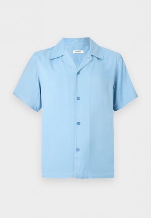 Άνδρας που στέκεται με τα χέρια στις τσέπες, φοράει ανοιχτό μπλε κοντομάνικο πουκάμισο με κουμπιά, μαύρα σορτς και μαύρα σανδάλια χωρίς λουράκια σε λευκό φόντο.