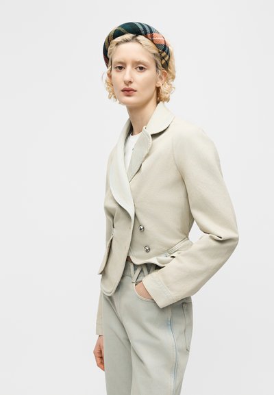 Νεαρός με κοντά σγουρά ξανθά μαλλιά, φοράει ένα μπεζ κοντό μπουφάν, ανοιχτόχρωμο παντελόνι και μια πολύχρωμη καρό κορδέλα στα μαλλιά, στέκεται με το ένα χέρι στην τσέπη.