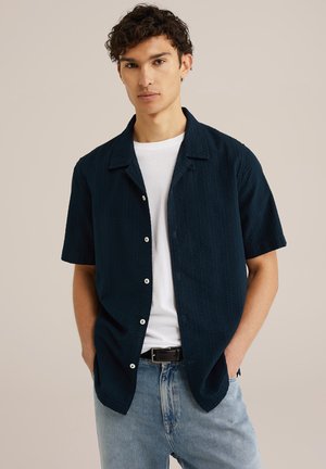Camisa de botones de manga corta en un tejido texturizado de color azul marino oscuro, llevada sobre una camiseta blanca, combinada con jeans de color azul claro. Detalles de botones blancos visibles.