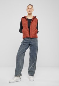 Quilted, rostfarbener Weste mit Kapuze und Reißverschluss, getragen über einem schwarzen Langarmshirt und kombiniert mit locker sitzenden blauen Jeans. Weiße Sneakers.