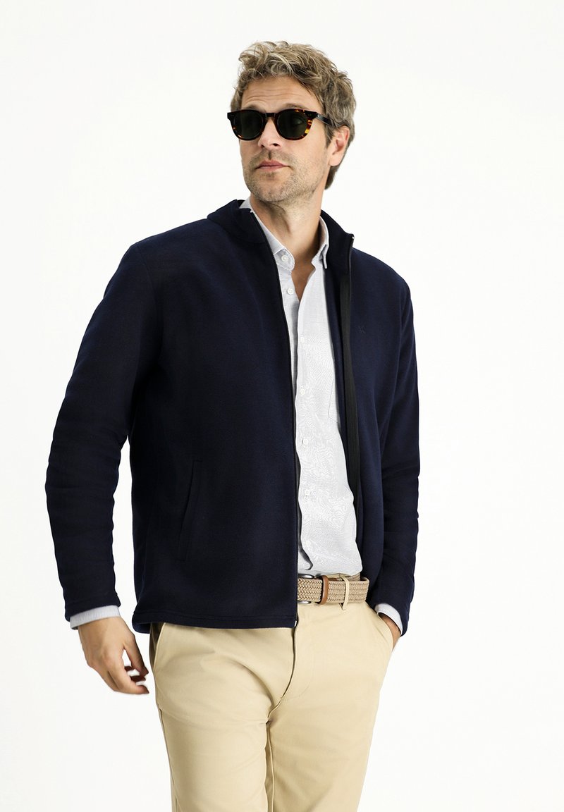 Granatowa polarowa kurtka z zamkiem z przodu, kieszeniami po bokach i wysokim kołnierzem. Nosiwana na jasnej koszuli w wzory i kremowych spodniach, dopełniona okularami przeciwsłonecznymi.