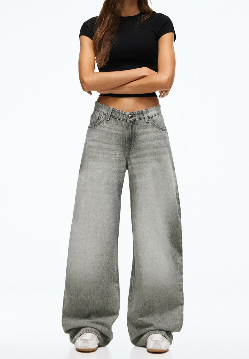 Femme debout, bras croisés, portant un crop top noir, un jean large gris clair et des baskets blanches sur un fond uni.