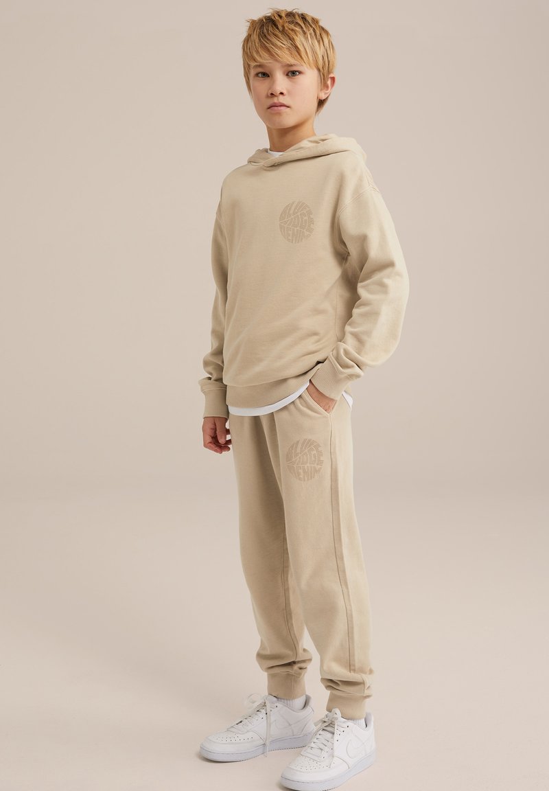 Beiger Kapuzenpulli und Sweatpants-Set mit passendem Logodruck. Baumwollmaterial, lockere Passform, gerippte Bündchen und kombiniert mit weißen Sneakers.