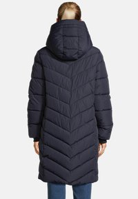 Frau in einem langen navyblauen, gesteppten Daunenmantel mit Kapuze, von hinten gesehen, vor einem hellen, einfarbigen Hintergrund stehend.