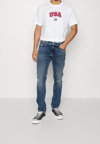 Vit t-shirt med röd "USA" grafik, kombinerad med blå denimjeans med slitna detaljer. Modellen har på sig svart och vita sneakers.