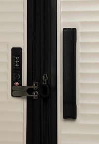 Primo piano di una valigia rigida beige con una cerniera nera, una serratura a combinazione e una robusta maniglia laterale nera.