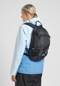 Sac à dos noir avec des côtés en maille et plusieurs compartiments, porté sur une veste zippée bleu clair. Dispose de bretelles ajustables et d'une poignée supérieure.