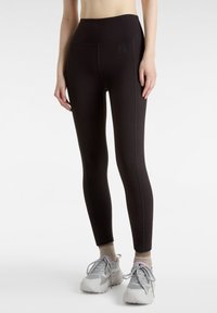 Höga svarta leggings i stretchmaterial, med platta sömmar och en liten logotyp. Bärs med grå sportskor och beiga strumpor.