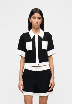 Schwarz-weißes kurzärmeliges Crop-Top mit Kragen, zwei Vorderentaschen und Knopfleiste. Kombiniert mit passenden schwarzen Shorts.