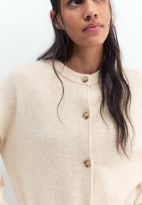 Tânără cu păr lung și închis la culoare poartă un cardigan crem cu nasturi, pe un fundal simplu, deschis la culoare.